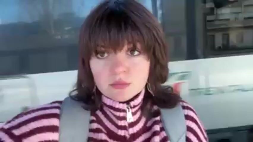 A young woman with brown hair and bangs, wearing a pink and maroon striped zip-up sweater and a gray backpack, stands in front of a bus or train. She looks directly at the camera and then turns her head to the right as the bus/train begins to move. The background shows reflections of buildings and trees. The environment appears to be a bus or train station.