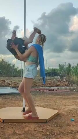 The video shows two women practicing pole dancing outdoors. The woman on the pole is performing a complex move where she is inverted and suspended horizontally by her hands and legs. The other woman is spotting her, providing support and guidance. The setting appears to be a rural or outdoor area with a dusty ground, some vegetation, and a fence in the background. The sky is cloudy.