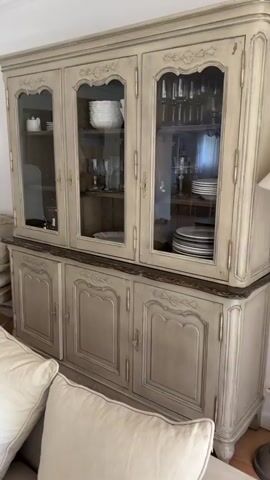 The video shows a brief, shaky pan of a living room, featuring light-colored walls, terracotta tile floors, and a neutral-toned sofa littered with pillows, a laptop playing a video, a purse, a bottle of water, and a striped throw blanket. The furniture is primarily light-colored and appears to be of a French country or farmhouse style.  A large china cabinet is visible, along with a dresser with a mirror, a writing desk, lamps, pictures on the walls, and an archway leading to another room, possibly a kitchen. Natural light streams in through a window covered with a sheer white curtain. The overall impression is a bright, casually decorated space.