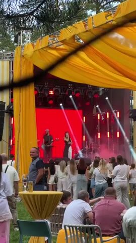 The video shows an outdoor concert venue with a yellow-draped stage and a red backdrop. A band is performing on stage, with a male guitarist on the left and a female singer in the center. A drummer is visible behind them. A crowd of people is gathered in front of the stage, some standing and some seated at tables. The environment appears to be a park or garden, with trees visible in the background. The lighting is a mix of stage lights and natural daylight.
