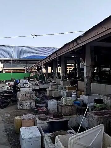 The video shows a marketplace with many boxes and containers stacked in front of a covered stall area. The boxes are made of various materials, including white plastic and some appear to be coolers. Many of them have writing on them, some illegible. There are also baskets, buckets, and other items scattered around the boxes. In the background, the covered stall area appears to be empty or sparsely populated, with a few people visible within. The ground is dirt or gravel. The overall impression is a fish market or a similar type of market that is either closed, preparing to open, or has just closed for the day.