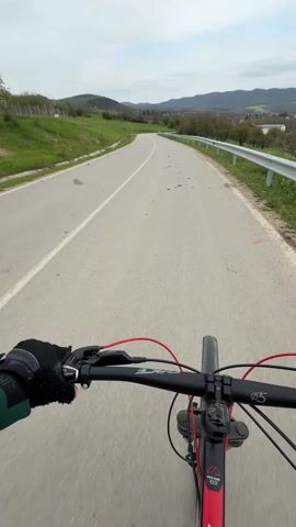 The video, shot from the first-person perspective of a cyclist, shows a road with a white line dividing the lanes and a silver guardrail on the right. The cyclist wears a black glove on their left hand and rides a mountain bike with red accents. Another cyclist, wearing black clothing, passes by on the left. The road curves gently, leading into a valley with green fields, trees, and mountains in the background under a cloudy sky. There are dark stains on the road surface.