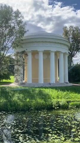 A circular white building with columns and a dome stands on a grassy hill next to a body of water covered in lily pads. Trees surround the building, and two people are walking in the distance on the grass. The sky is blue with white clouds.