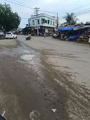 The video shows a street scene with several stalls lining the right side of the road, displaying fruits and other goods. A white building with a green trim is visible on the left, with power lines running overhead. Several motorbikes and a truck are present on the road. The road surface appears to be asphalt and is uneven with some puddles and debris. The sky is overcast. People are seen at the stalls and riding the motorbikes. One stall has a sign that reads "AYAN SERVICE HP". The scene appears to be a market area in a town or city.