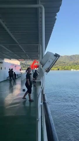 A video opens with a view from the deck of a ship. The perspective is looking outward from the starboard side, with the deck extending to the left and the ocean to the right. A white railing runs along the edge of the deck. A large, grey light fixture is mounted on a white pole on the right side of the frame. The deck is green and appears to be wet in some areas. In the background, on the left side of the deck, there are several people walking and standing. One woman in a black dress is prominent in the foreground, facing away from the camera and looking out at the water. Further down the deck, a red lifeboat or life-saving equipment is visible. Beyond the ship, the ocean is calm and reflects the clear blue sky. In the distance, a green, hilly coastline with scattered trees and some structures can be seen. The sun is shining, casting shadows on the deck. The video is very short, lasting only a few seconds, and shows a static or slow-moving scene.