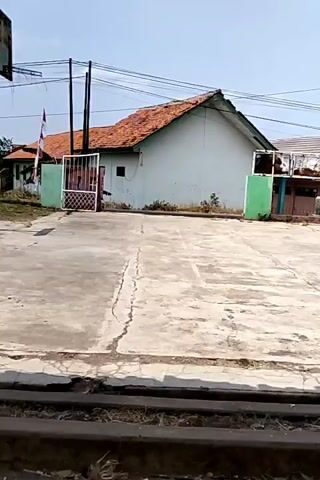 The video captures a concrete area in front of a light blue building with a reddish-brown tiled roof. The building has a white window and a white door. There are two gates, one on the left and one on the right. The gate on the left is white and appears to be made of metal bars. A flag on a pole stands behind the left gate. The gate on the right is green. In the middle of the video, a silver van enters the area through the left gate and parks in front of the building. The van has some writing on its side. The van then exits the area through the left gate. The concrete area is cracked in several places. In the foreground, there are horizontal dark lines that appear to be railway tracks or some similar structure. The sky is blue and clear.