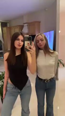 Two young women are posing for a selfie in what appears to be a modern apartment. The woman on the left has long, dark hair and is wearing a black top and blue jeans. She is holding the phone and making a kissing face. The woman on the right has blonde hair with dark roots, a white long-sleeved zip-up shirt, and blue jeans with a belt. She is also making a kissing face. The background shows a clean and well-lit apartment with a kitchen area, a large television displaying colorful images, and potted plants. The floor is shiny and reflective. There is music playing in the background.