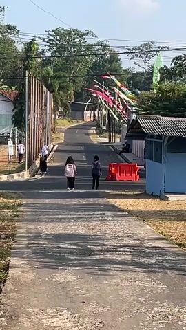 A motorcycle with a sidecar is approaching a gate on a paved road. Several people, including two individuals wearing hijabs and one wearing a blue jacket, are walking on the road ahead of the motorcycle. The motorcycle is being ridden by a person wearing a helmet and a brown shirt. There are orange barriers on the right side of the road. The environment appears to be outdoors with trees and buildings visible in the background. Colorful flags are strung across the road in the distance.