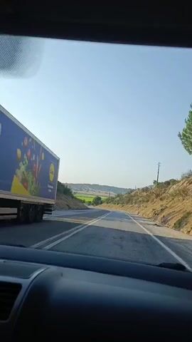 The video shows a first-person perspective from inside a car driving along a paved road. The road has clear lane markings and appears to be in a rural or semi-rural area. The landscape consists of dry, grassy hillsides, some sparse vegetation, and trees. The sky is clear and blue. There are some utility poles visible along the roadside. In some segments, the view through the windshield shows a bright sky with some light reflections or streaks. The camera occasionally pans or the car turns, providing views of the surrounding terrain and the interior of the car, including parts of the dashboard, windshield, and side mirror. The lighting suggests it is daytime, likely morning or afternoon. There are no other vehicles or pedestrians clearly visible on the road.