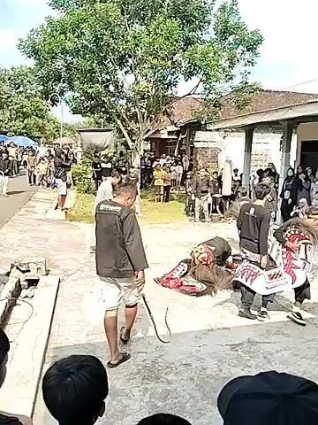 The video shows an outdoor cultural performance. A group of people are gathered to watch, and several performers are in the center of a paved area. One performer is on the ground, appearing to be in a theatrical pose, wearing a costume that resembles an animal with a long, shaggy mane. Another performer, dressed in black and white, stands over the prone figure, holding a whip. A man in a black jacket and white shorts, also holding a whip, stands to the left, facing the performers. In the background, more performers in traditional costumes are visible, and a crowd of spectators lines the sides of the area. Buildings and trees form the backdrop of the scene. The atmosphere appears to be festive.