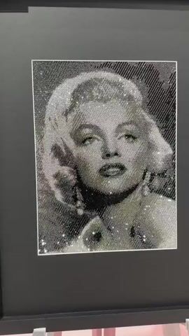 The video shows a close-up, slightly tilted shot of a framed artwork. The artwork is a portrait of Marilyn Monroe, rendered in what appears to be diamond art or a similar mosaic technique using small, reflective beads or crystals. The portrait is in black and white, with variations in the density and color of the beads creating shading and detail. Marilyn Monroe's face is centered in the frame, with her hair styled around her head and shoulders. She is looking directly at the camera with her mouth slightly open. The artwork is matted with a dark border and enclosed in a black frame. The background behind the artwork is a plain dark surface. The lighting highlights the reflective quality of the beads, making the portrait shimmer. The video appears to be taken in a gallery or exhibition setting.