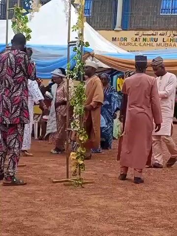 The video shows an outdoor gathering with several people on a red dirt ground. In the foreground, two metal arch-like structures adorned with artificial yellow and green flowers frame a central area where individuals are dancing. A man in a patterned red, black, and white outfit on the far left films with a phone. To his right, a woman in a white lace dress dances. Next to her, a man in a brown traditional robe moves. Further right, a woman in a sparkling gold outfit dances, and behind her, another woman in a blue patterned dress stands. On the far right, a man in a light pink traditional outfit dances, while another man in a darker traditional outfit walks away from the center. In the background, white and blue tents are set up, and a banner with Arabic text and "MADR SAT NURU LAH" is visible on a building. Cars are partially visible behind the tents.