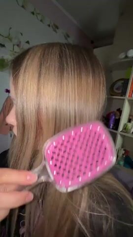 A woman with long, blonde, straight hair brushes her hair with a pink brush. Her hair appears healthy. The woman is wearing a black shirt. A shelf with books and other items is in the background, as is a tiled wall.