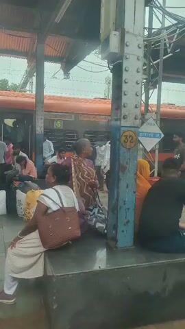 This video shows a busy train platform. Several people are visible, some sitting on the platform with luggage, others standing or walking. There is a train at the platform, with its doors open. One person appears to be operating a floor cleaning machine. The platform is covered by a roof, and there are metal support columns. The ground is paved and wet in some areas. The overall atmosphere is one of transit and activity at a public transportation hub.