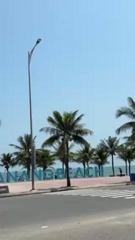 The video shows a roadside view of Da Nang Beach, Vietnam. Palm trees line the road, partially obscuring the large, turquoise "DA NANG BEACH" sign. A street lamp stands tall, next to the sign. A car drives by and a motorbike is parked near the sidewalk. In the background, there is the ocean. The sky is clear and blue.