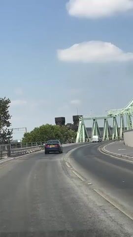 The video shows a view from a moving vehicle driving on a road. In the foreground, the asphalt road is visible with yellow lane markings. A dark-colored car is driving ahead on the road. In the background, a large green bridge with a distinctive arched structure dominates the scene. The bridge is a prominent truss bridge, with its green metalwork clearly visible against the sky. Beyond the bridge, there are trees and what appears to be a historical building with towers, possibly a castle or a fortified structure, which is a landmark in Runcorn, United Kingdom. The sky is blue with a few white clouds, suggesting a bright, clear day. The overall scene depicts a journey across a bridge in an urban or semi-urban environment. The time of day appears to be daytime, possibly late morning or afternoon, given the bright sunlight and clear sky. No specific emotions are conveyed, and the atmosphere is neutral, focusing on the visual depiction of the location and the bridge. There is no visible text within the frame.