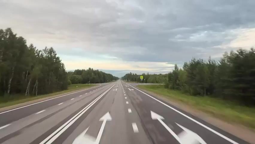 The video shows a first-person perspective of a car driving on a two-lane highway. The road is wet, indicating recent rain. Both sides of the highway are lined with dense green forest. The sky is overcast with dramatic clouds. Traffic lanes are marked with white lines, and there are directional arrows painted on the road. A signpost is visible on the right side of the road. The car appears to be traveling at a moderate speed.