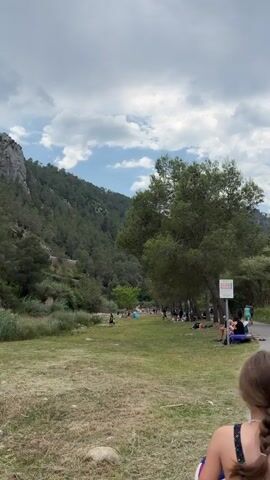 The video shows a park area near a mountain on a partially cloudy day. People are gathered on the grass, some sitting under trees, others walking around. A paved path runs along the edge of the park area. There are trees scattered throughout the park and densely covering the mountainside. A sign with information is posted near the path. The overall scene suggests a relaxed, outdoor recreational environment.