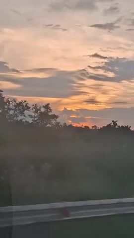 The video shows a time-lapse of a sunset over a rural landscape. The sky is filled with soft, orange and pink clouds as the sun dips below the horizon. In the foreground, there is a blurry view of trees and a road. The overall mood is peaceful and serene.