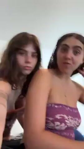 Two young women are filming themselves indoors. They’re wearing tube tops. They’re lip-syncing and dancing to music. One is wearing a purple and white tube top, the other is wearing a brown one. They put their arms around each other and make hand gestures at the camera. The environment appears to be a bedroom or apartment. There is white paint on the wall.