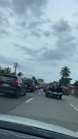The video is taken from inside a car driving on a highway in the Philippines. The traffic is slow-moving due to congestion. Various vehicles are visible, including a black SUV, pickup trucks, a bus, and a tricycle. The environment is tropical, with palm trees visible along the road. On the side of the road, there is a small shop with motorbikes parked in front of it, and a basket of durians. The sky is cloudy, suggesting overcast weather.