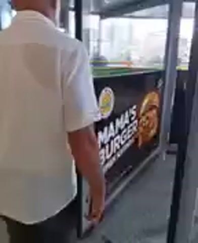 A man in a white shirt walks away from the camera. He is near a storefront with a sign that says "MAMA'S BURGER". Other people are visible in the background, walking outside.