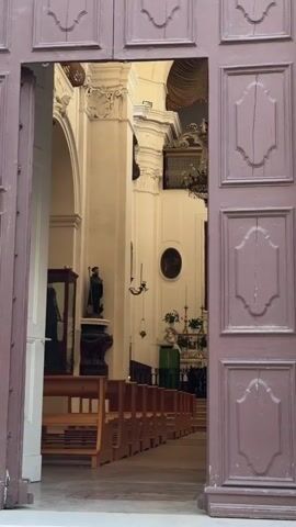 The video shows the interior of a church, viewed from an open doorway framed by dark wooden doors. The camera is static.  Inside the church, rows of wooden pews are visible, leading towards an altar area.  The altar is adorned with religious symbols and decorations, including a large painting above it. A chandelier hangs from the ceiling.  To the left of the pews, a statue of a robed figure holding a staff is visible.  On the right side of the pews, a person is seen kneeling or bending over, partially obscured by the pews.  The church architecture features high ceilings, columns, and ornate decorative elements.  A poster or sign is attached to the left door frame.