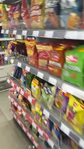 The video shows a tour through the aisles of a convenience store or small supermarket. The camera moves past shelves stocked with snack chips, drinks, and refrigerated items such as yogurt, pre-made salads, meats, and cheeses. The lighting is bright and the shelves are well-stocked. No people are visible in the frame.