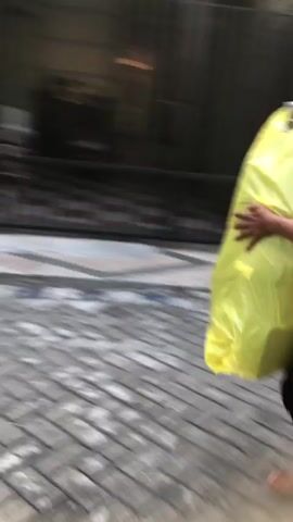 The video is extremely blurry and shaky, making it difficult to discern many details. It appears to be an outdoor scene. A person, whose body is mostly obscured by motion blur, is holding a large, bright yellow bag. The person's arm and hand are visible, gripping the top of the bag. The background is also heavily blurred, but suggests a paved area, possibly a sidewalk or street, with some darker structures behind it. The person and the bag are in motion, moving from left to right across the frame. The lighting appears to be natural daylight. The overall impression is one of hurried movement in an urban or semi-urban setting.