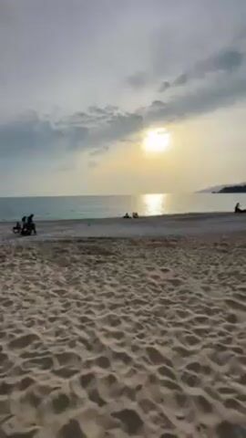 The video shows a beach at sunset. The sky is cloudy, but the sun is still shining brightly. The beach is sandy and there are a few people scattered along the shoreline. In the distance, there are some mountains. At the end of the video, a woman smiles at the camera. She is holding a baby carrier.