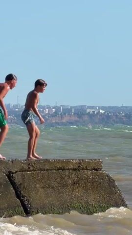 Two young boys are on a concrete pier. The boy on the left is wearing green swim trunks, and the boy on the right is wearing blue and white patterned swim trunks. The boy on the left jumps off the pier into the choppy, brown water. The boy on the right follows, with his arms extended as he jumps. He lands in the water, creating a large splash. The younger boy emerges from the water and swims. In the background, there is a distant city skyline and a bright orange buoy. The sky is clear and blue.