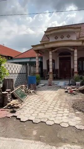 The video shows an outdoor scene, likely a residential area undergoing some form of construction or renovation. In the foreground, there is a partially paved driveway or walkway made of hexagonal concrete tiles. Some of these tiles are neatly laid, while others are stacked or scattered, indicating ongoing work. Debris, including rubble, gravel, and broken concrete, is visible in several areas.

To the left, a wooden structure, possibly a planter or a base for something, stands near a pile of bricks and a green chalkboard propped against it. Further back, a blue barrel is visible. Beyond this, there's a white wall with a patterned design, and behind it, a house with a red tiled roof.

To the right, the front of a house is partially visible. It has a porch supported by columns and a brown double door. Some building materials and tools are present near the entrance. Trees and foliage are also present, suggesting it is daytime. The sky is partly cloudy. There are no people visible in the frame. The overall impression is of a work-in-progress at a property.