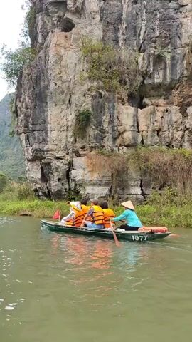 The video shows a small boat with four people paddling down a river lined with lush greenery and tall rock formations. The people are wearing life vests and one person in the back is wearing a conical hat. The boat is green with the number 1977 painted on the side. The water is calm and greenish in color. Other boats with similar configurations can be seen in the distance on the river. The sky is overcast.