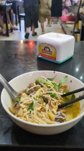 The video opens with a close-up of a bowl of noodles, chopsticks, and a spoon on a black table. A tissue box with the "MEIZAN" logo is also visible. The camera then slowly pans up and back to reveal the interior of what appears to be a small restaurant. Several patrons are present, some seated at tables, while others are standing near the entrance looking at food displays. Red lanterns hang from the ceiling. The restaurant is open to the street, allowing a view of the buildings across the road. Some of the people inside are wearing face masks.