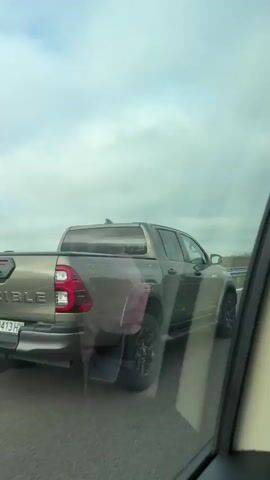 The video shows a view from inside a car driving on a highway, looking out the passenger side window. Initially, a tan Toyota Hilux pickup truck is visible in the lane to the right, with a license plate A 8413 HX and the word "INVINCIBLE" on the tailgate. The camera then follows the road as the car continues to move, showing the roadside with a metal guardrail, fields, scattered trees, and power lines in the background under a cloudy sky. The view is somewhat obscured by reflections in the car window.