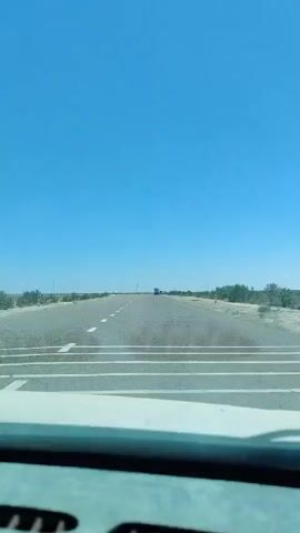 The video shows a view from inside a vehicle looking out through the windshield at a straight, paved road. The road appears to be in a dry, arid environment with sparse vegetation on either side. The sky is a clear, bright blue, indicating a sunny day. There are white dashed lines on the road, marking the lanes. In the distance, a few vehicles, possibly trucks, can be seen traveling on the road. The dashboard of the vehicle is partially visible at the bottom of the frame. The overall impression is of a long, open road journey through a desolate landscape.
