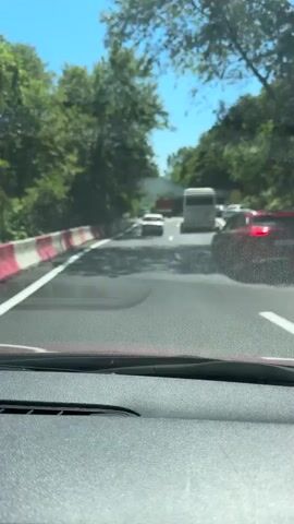 The video is filmed from the perspective of a passenger inside a vehicle, looking forward. The vehicle is on a road with multiple lanes. In front of the vehicle, there are other vehicles including a white car, a large white bus, and a red SUV. There are trees and foliage on both sides of the road, suggesting a scenic route. The sky is clear and blue, indicating a sunny day. The road surface appears to have some unevenness or patches. The vehicle is moving at a moderate speed, as indicated by the slight blur of the surrounding environment. The traffic appears to be moving smoothly, with no obvious signs of a standstill or accident. The barrier on the left side of the road is made of white and red plastic.
