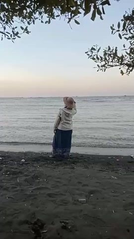 A woman wearing a hijab and a hat stands on a black sand beach facing the ocean. She is wearing a light-colored jacket over a long blue skirt. The ocean is calm, with gentle waves lapping at the shore. The sky is a pale blue, with a hint of pink near the horizon, suggesting it is either sunrise or sunset. Trees with green leaves frame the top of the shot, casting shadows on the scene. The overall mood is peaceful and serene.