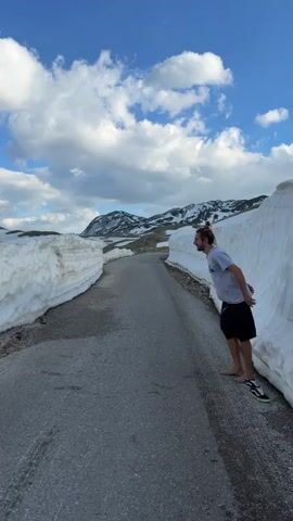 A man with long hair in a bun, wearing a grey t-shirt and black shorts, is barefoot on a paved road between high snowbanks.  He jumps onto the snowbank, landing on top, and then tests the surface with his feet. The background shows a partly cloudy blue sky and snow-covered mountains.