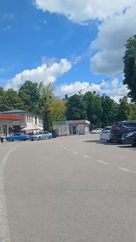 The video shows a street scene on a sunny day with a bright blue sky and scattered clouds. In the foreground, there is an asphalt road with white markings. A row of buildings runs along the opposite side of the street. The buildings appear to be commercial establishments, with some displaying signs. One sign reads "КОПІЙКА" which is a Ukrainian supermarket chain, and another displays "АПТЕКА" which means pharmacy. Several cars are parked in front of the buildings, including a blue vintage car, a silver sedan, and a white SUV. There are also a few people walking around and sitting outside. In the distance, trees and greenery are visible, indicating a somewhat suburban or town setting. The camera pans slowly from left to right, capturing different parts of the street.