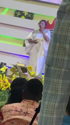 A woman in a shiny, white gown is singing into a microphone on a brightly lit stage. She is standing behind a podium decorated with yellow flowers and green foliage. Behind her, the stage is tiered and lit with green and blue lights. Several people sit in the foreground facing her; one man is wearing a pink shirt with red patterns, and another man stands off to the side, wearing a blue and grey plaid suit.