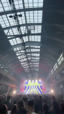 The video shows a large crowd of people at what appears to be an indoor concert or rave. The venue is a spacious industrial-looking building with a high ceiling made of glass panels. Overhead are visible heavy-duty rigging and lighting equipment. At the far end of the hall is a large stage with a bright LED screen displaying colorful visuals, mostly yellow and blue. Stage lights are active, adding to the atmosphere. The crowd is dense, with people facing the stage. The overall mood is energetic and lively.