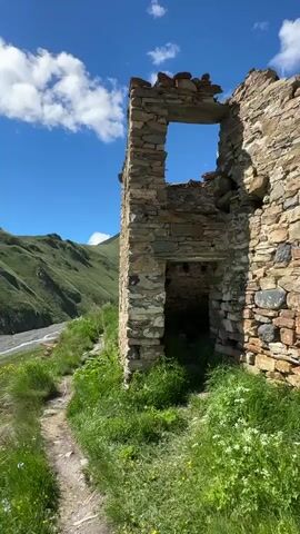 The video opens on a close-up of a stone ruin bathed in sunlight, with green grass and wildflowers growing around it and a dirt path leading away. The view then pans to reveal a vast mountain landscape under a bright blue sky with scattered clouds. In the foreground, lush green hills roll down to a wide, rocky riverbed, with a river flowing through it. Further in the distance, more dramatic, snow-capped mountains rise, their peaks shrouded in clouds. The sun casts a bright lens flare across the sky. The camera continues to sweep across the panorama, showcasing the expansive valley, power lines, and more distant, jagged peaks. The final shot captures a large, green mountain with patches of snow near its summit, framed by the bright sky and foreground foliage. The overall scene depicts a remote and wild mountainous environment on a clear, sunny day.