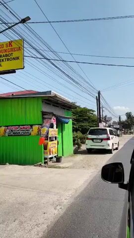 The video shows a view from inside a car traveling down a street. On the left side of the street, there are buildings with corrugated metal siding and signs, including one that says "ANEKA". There are also utility poles with many wires running across the street and over the buildings. A white van is parked on the side of the road. The road is paved in some areas and unpaved in others. The sky is clear and blue. The car appears to be moving at a moderate speed.