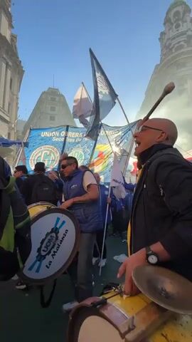 The video shows a demonstration or rally in a city, with multiple individuals marching down a street. The camera is positioned among them, focusing on two men beating large drums. The drums are labeled "UOM, La Matanza." The backdrop includes a banner reading "UNION OBRERA METALURGICA," and flags are waved overhead. Smoke fills the air. People surrounding the drummers are seen raising their arms in apparent support. The architecture in the background consists of tall, ornate buildings under a blue sky.