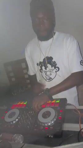 A DJ, wearing a white t-shirt with a cartoon graphic and a brown baseball cap, is seen operating a DJ controller. The DJ controller has two illuminated platters and various knobs and sliders. Next to the controller, there are two microphones. In the background, another person, wearing a black and white shirt, is holding a microphone to their mouth and giving a thumbs-up gesture. The environment appears to be a dimly lit indoor space, possibly a party or event, with a smoky atmosphere.