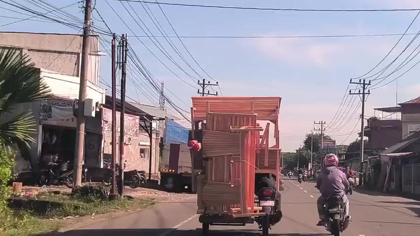 The video depicts a street scene with a motorcycle pulling a large, unstable load of what appears to be wooden furniture frames. The motorcycle is moving along a paved road with a few other motorcycles and a truck alongside. There are buildings and utility poles lining the side of the road. The sky is visible, a light blue with few clouds. The focus of the video is clearly the motorcycle with the precarious load.