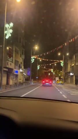 The video shows a night drive through a city street. The view is from inside a car, looking out through the windshield. The road is lined with buildings, streetlights and decorations, like Turkish flags and illuminated flower designs strung between lampposts. A red car is in front of the vehicle, driving in the same direction. A yellow taxi is parked on the right side of the road with a few people standing nearby. The audio features music with a strong beat.