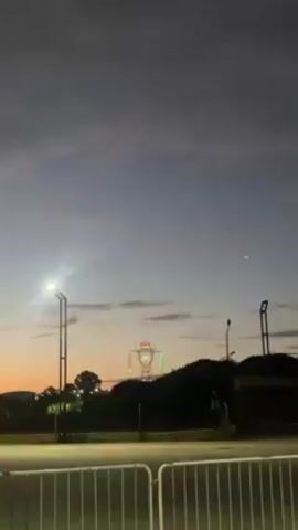 The video shows a power line tower decorated with lights, creating the appearance of a robot-like figure. The tower stands behind a row of trees on a slight hill, with a sunset in the background. A street light illuminates a parking area with a white metal fence in the foreground. The sky is filled with soft clouds, and the light is dim. There are no people visible in the video.