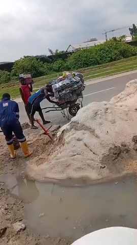 The video shows people working on a construction site near a road. Several men are shoveling sand and moving it in wheelbarrows. One of the wheelbarrows is heavily loaded with stacks of what appear to be packaged goods. The workers are dressed in work clothes, including a blue jumpsuit and yellow boots. There is a puddle of water on the ground, and the construction area is muddy. In the background, there is green vegetation, a road, and some buildings. Towards the end of the video, the camera shows a constructed form with rebar, presumably to be filled with cement.